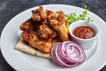 Grilled chicken wings with barbecue sauce, pita bread, microgreen and onion rings, on a white plate, against a dark background