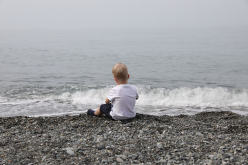 A little boy with blond hair sits on a pebble by the sea.