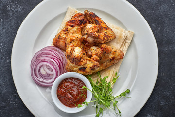 Grilled chicken wings with barbecue sauce, pita bread, microgreen and onion rings, on a white plate, against a dark background. Top view