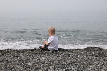 A little boy with blond hair sits on a pebble by the sea. The boy is wearing a white T-shirt. Cloudy weather.