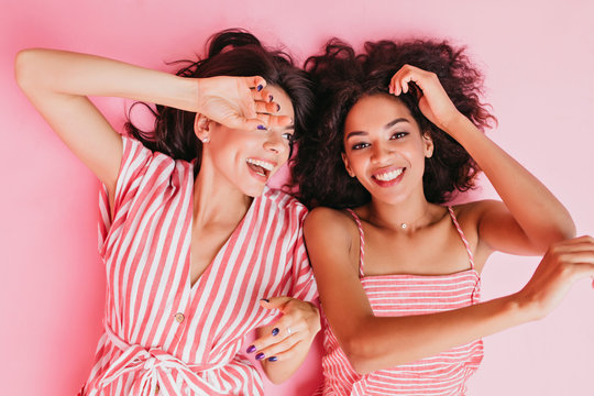 Cousins in wonderful mood are lying on floor in high spirits. Girls pose with sincere smiles in pink clothes for close-up portrait
