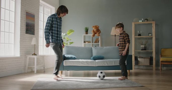Happy father and son playing football together at home. Caucasian man dribbling the ball while young son tries to take it from him - active games, happy family concept 4k footage