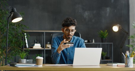 Arab young male office worker in glasses sitting at desk, holding credit card and shopping online on laptop computer.