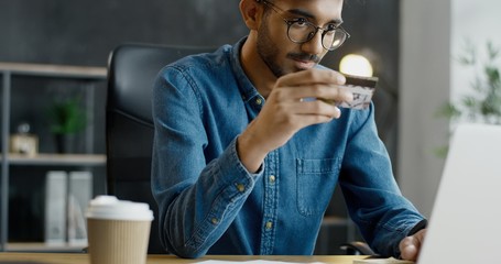 Arab young male office worker in glasses sitting at table, holding credit card and shopping online...