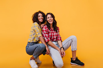 American women in trendy street style clothes pose sitting on floor in orange studio. Photo indoors of young girls laughing sincerely