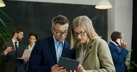 Caucasian male and female business partners standing at hall of conference and watching on tablet device while tapping and scrolling.