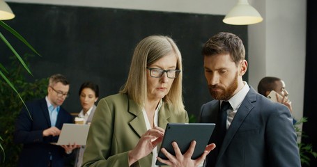 Female boss with male company worker standing together in hall at break of conference and watching video on tablet.