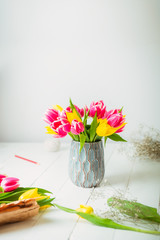 Fresh spring yellow and pink tulips bouquet in a vase, other flowers and details on florist workspace on white wooden table. Education of floristry. Selective focus. Vertical card. Copy space.