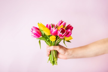 Male hand holding beautiful fresh pink and yellow tulips bouquet on light pink background. Greeting card mockup. Birthday, mother's and woman's day. Selective focus. Copy space.