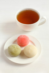 Three types of Japanese dessert mochi - pomegranate with honey, green matcha tea, coconut on a white saucer and a cup with tea on a white wooden table.