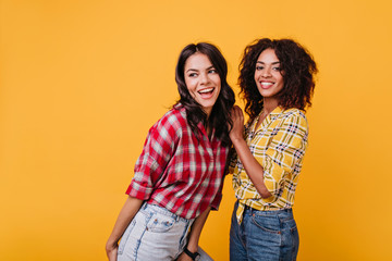 Happy young women dancing on yellow background. Indoors portrait of two stylish girls in jeans