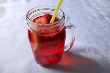 Strawberry smoothie chopped into slices and a slice of lemon in a glass jar decorated with a drinking tube on a home desk. Summer and season food and drink concept. Close up, selective focus