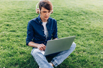 Portrait of a beautiful young man sitting on the grass in a Park with a laptop and listening to music with headphones