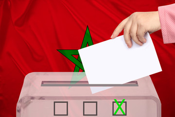 female voter drops a ballot in a transparent ballot box on the background of the Morocco national flag, concept of state elections, referendum
