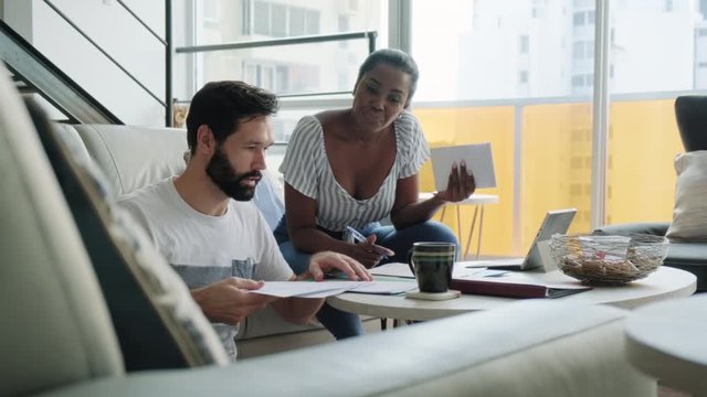 Interracial Couple Doing Tax Returns And Preparing Home Budget In Living Room. Black Wife And White Husband Checking Utility Bills And Bank Account. Married People Preparing Taxes And Forms