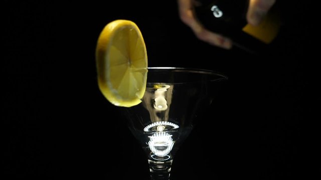 A man pours a martini (white wine) from a bottle into a glass goblet decorated with a slice of lemon on a black background. Closeup. The camera moves around. Slow motion. High speed camera
