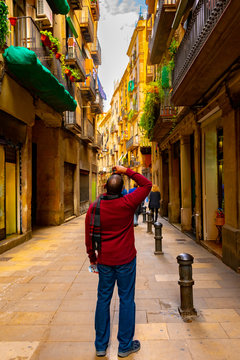 Male tourist stops in middle of narrow backstreet in Barcelona to take photograph of high rise apartment building balconies with railings and hanging plants.