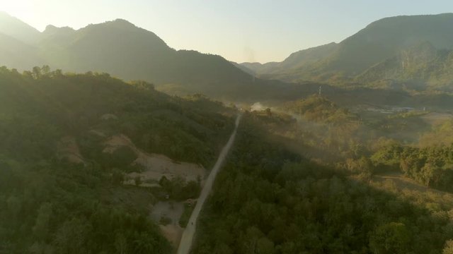 Aerial shot of road amidst green trees near village against sky, drone ascending over landscape during sunrise - Tat Sae Waterfalls, Laos