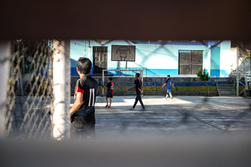 Fototapeta premium View on the group of young boys playing football at the concrete playground at the school campus behind the fence.