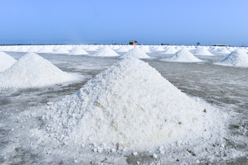 White sea salt is scooped up into piles like mountains and dried in the sun. Behind is a clear blue...