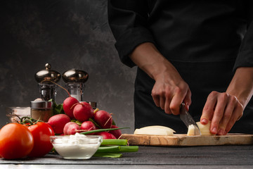 The chef cuts potatoes for cooking potatoes in French. Against the background of vegetables. With space for advertising, restaurant business, and cooking. Book of recipes