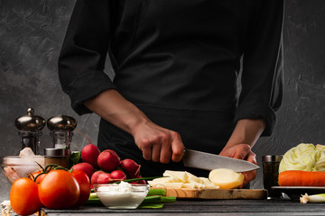 The chef cuts potatoes for cooking potatoes in French. Against the background of vegetables. With space for advertising, restaurant business, and cooking. Book of recipes