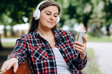 Cute woman, plus size, sitting on a bench in headphones, looking at a smartphone