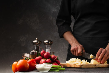 Chef cooks a dish with potatoes, slicing and cystitis. Against the background of vegetables, preparing French fries. Restaurant business, tasty and wholesome food