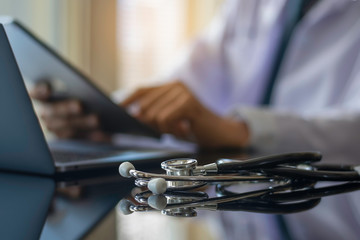 Male doctor in white lab coat hand holding and using modern digital tablet, work on laptop computer with  stethoscope on the desk at workplace. Medic tech,e health, online medical or ehr concept. 