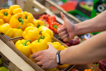 Close-up of mans hands with fitness tracker showing a thumb up approvingly selecting yellow peppers in the grocery stores fresh vegetable department