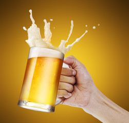 Hand holding a mug of light beer toasting with bubble froth splash in golden background with copy...