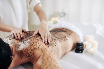 Cosmetologist applying coffee peeling on back of male client to exfoliate ot before massage