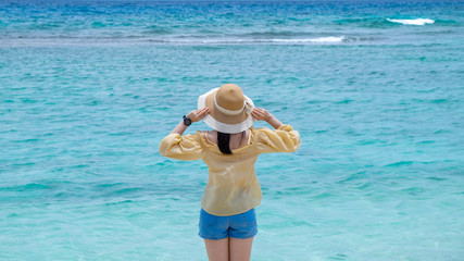 Young woman in yellow blouse and short blue jean with straw hat standing on the beach with blue sky that relaxing and enjoying on vacation.