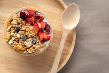 Top down view of yogurt with muesli, strawberry, cherry and dried raisins in a light brown wooden bowl next to spoon with copy space