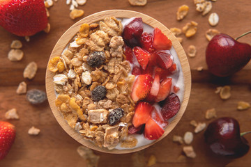 Close-up top down view yogurt with muesli, strawberry, cherry and dried raisins in a brown wooden bowl on wood background.