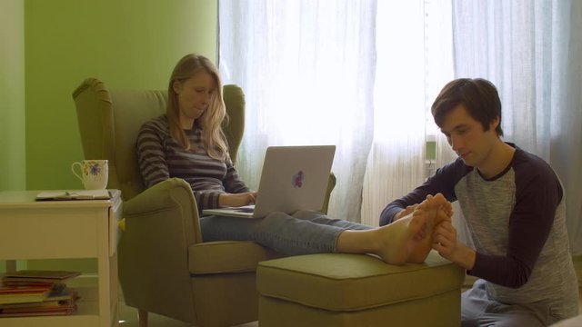 Young woman working remotely. She is sitting with laptop in the chair and drinking tea. Her husband doing her a feet massage. Daily family life, remote job.