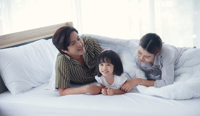 Happy young Asian family with a daughter are lying on bed under the blanket and Patting on daughter’s head in the bedroom. Dad mom little girl playing and spend time together