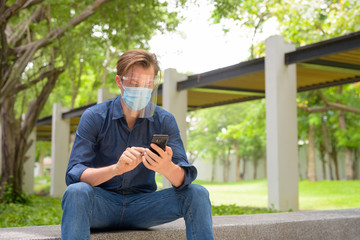 Fototapeta premium Young man using phone with mask and face shield while sitting at the park