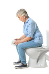 Elderly woman with paper sitting on toilet bowl against white background. Concept of hemorrhoids