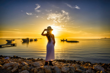 Woman in white dress wearing hat standing on the beach over sunlight seaview with boats on water and colorful sky background, Bahrain.