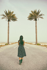 The back of woman wearing dress walking alone in the middle road with palm trees over clouds sky background, Bahrain.
