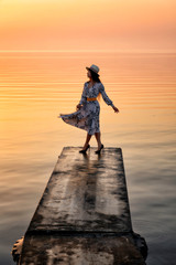 Motion blur of woman waring dress and hat dancing on the pier over colorful twilight sunrise morning sky over sea water background.