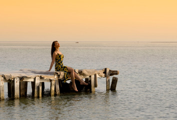 Beautiful woman sitting on wooden old pier on sea water relaxing over sunset orange sky background, Bahrain.