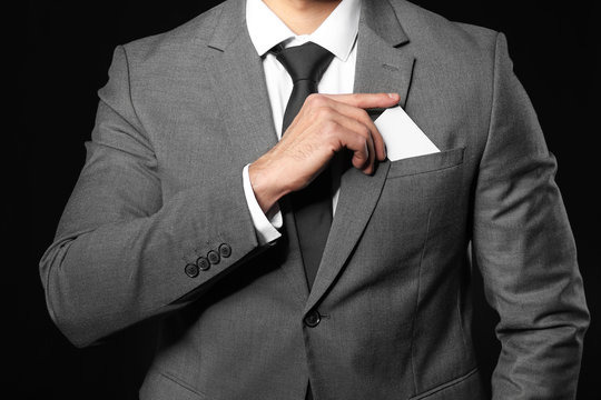 Handsome young man in elegant suit taking business card out of pocket on dark background
