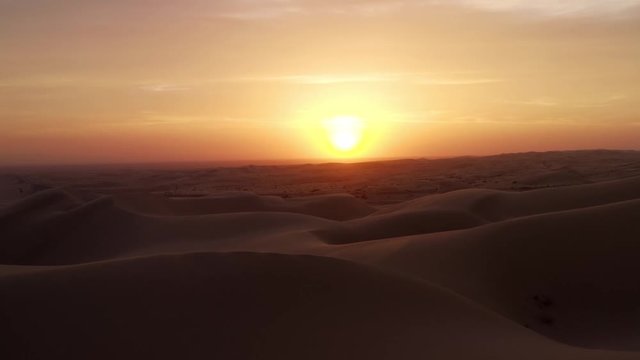 The camera pulls back over many sand dunes revealing a small figure below during a fiery sunset.