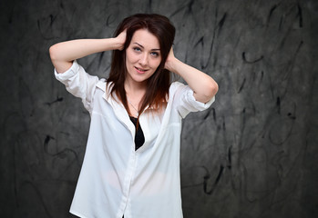 A pretty model is standing on a gray background in a white shirt. Studio portrait of a caucasian brunette girl with minimal makeup.
