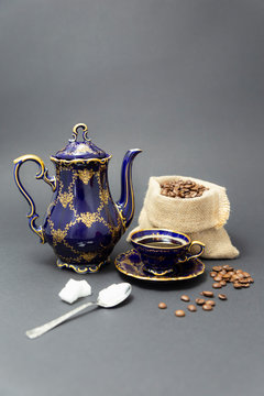 Still life with a beautiful cobalt blue colored vintage porcelain coffee set with golden floral pattern, a silver spoon with sugar cubes and a gunnysack filled with roasted coffee beans.