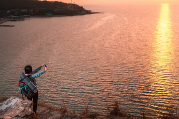 Fototapeta premium A woman is standing on the edge of a cliff and taking photos of the sunset on her smartphone. In the background, the sea at sunset. Copy space. Concept of Hiking and outdoor activities