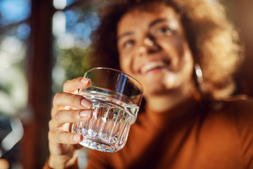 Closeup of mixed race girl sitting in cafe and holding glass of fresh water. Selective focus on glass.