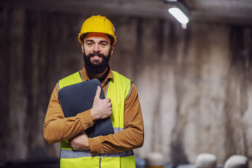 Young good-looking smiling bearded worker in vest with laptop in hands standing inside of tunnel...